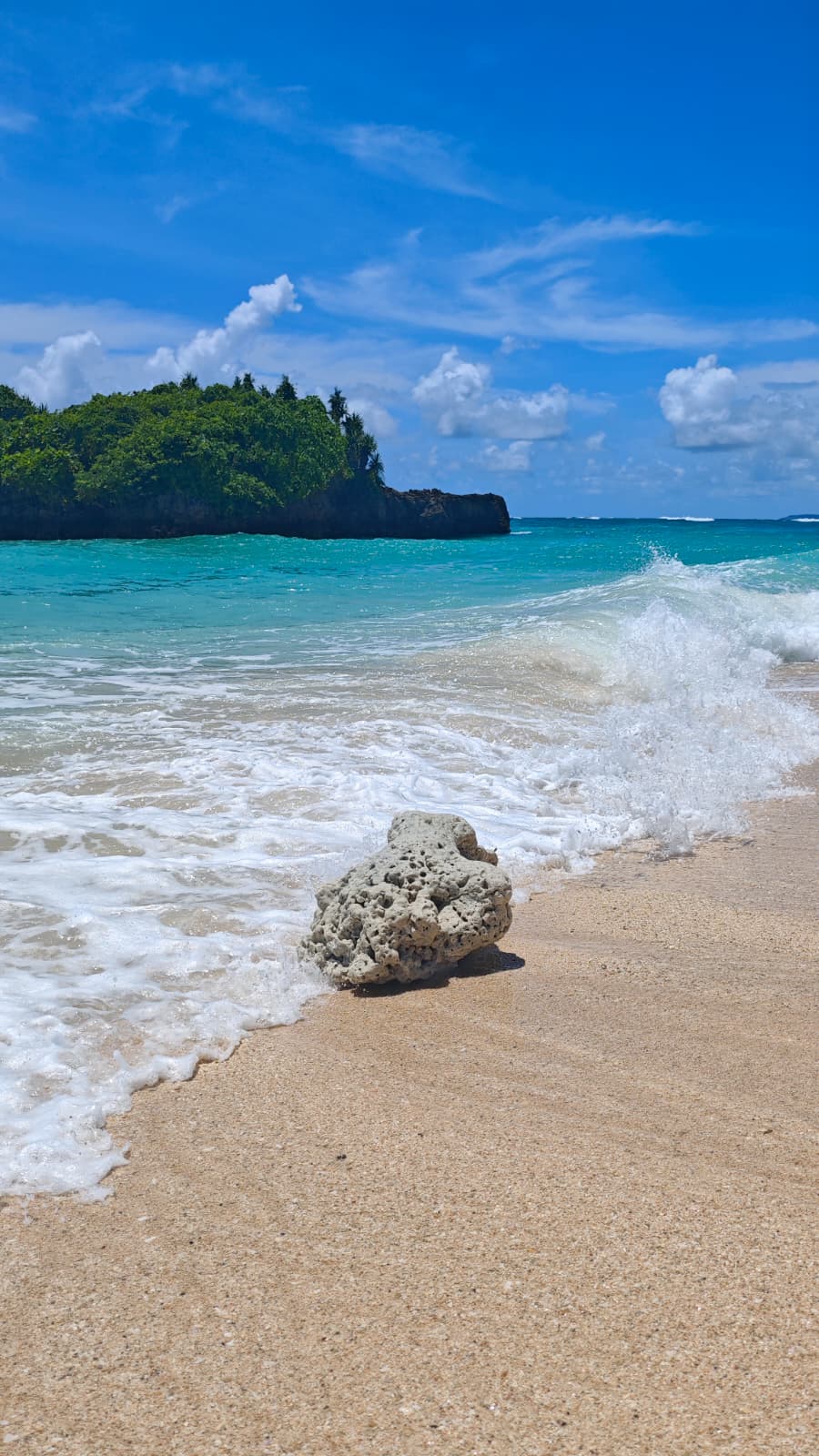 Vague qui vient lécher un rocher sur une plage de sable, face à une île